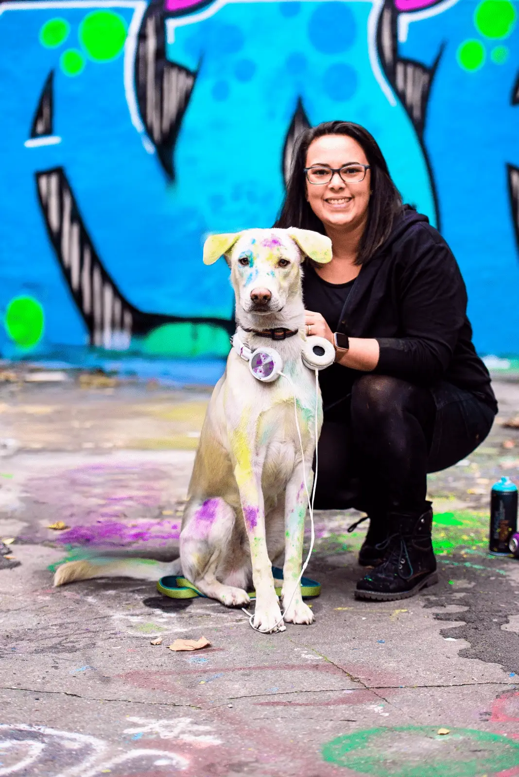 Frau in schwarzer Kleidung kniet lachend neben einem hellen Hund, der mit bunter Farbe gesprenkelt ist. Beide befinden sich vor einer leuchtend blauen Graffiti-Wand. Der Hund tr&auml;gt Kopfh&ouml;rer um den Hals und blickt ruhig in die Kamera.